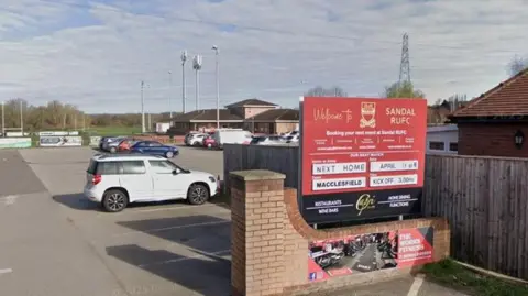 Entrance to Sandal RUFC on Standbridge Lane in Wakefield, with a sign bearing the club's name on the left, and a car park and rugby pitches in the background.