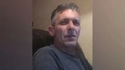 A man in his mid-fifties with grey hair. He is sitting in an armchair and squinting at the camera.