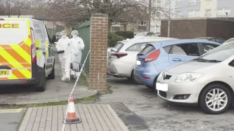 Malik Walton/BBC Two people wearing white forensic outfits walk near a police van and a cordon. Parked cars are also next to them by a green metal and brick fence. 