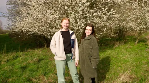 Two women stood in front of a blossom tree. The woman on the left has red hair with a cream jacket and black t-shirt with light green trousers. The woman on the right has long brown hair and is wearing a green full-length coat