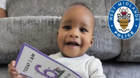Family/West Midlands Police Noah at six months old, in a cream jumper. He has tufts of curly black hair. He is holding a purple and white card that says "today I am six months old".