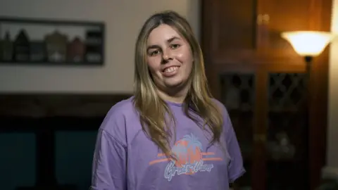 A young woman in a purple T-shirt looks into the camera