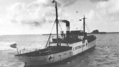 The SS Vega ship in the water in Guernsey's Saint Peter Port harbour in 1944. The white ship has two foldable masts with the red cross symbol painted on it in three locations.