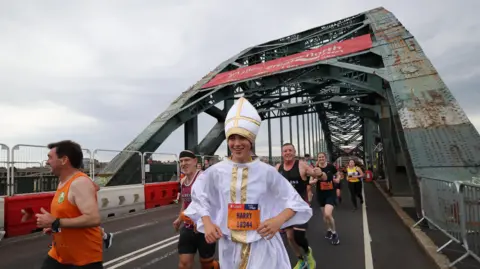 Young man dressed in pope costume runs across the Tyne Bridge. There are several runners on the bridge nearby. Everyone is smiling. 