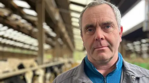 Man with grey hair looking into the camera. He is wearing a blue fleece and a grey jacket. He is in a large cow shed with blurred cows in the background.