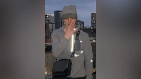 Greater Manchester Police A teenage boy wearing a hoodie and a grey beanie stands on a balcony with skyscrapers in the background. He is holding his stand up in front of this face making a gesture.
