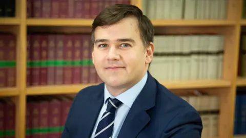 A middle aged man looks towards the camera with a neutral face. He has dark hair and is wearing a light blue shirt with a navy jacket and tie. He is in front of a bookshelf holding various leather bound books.