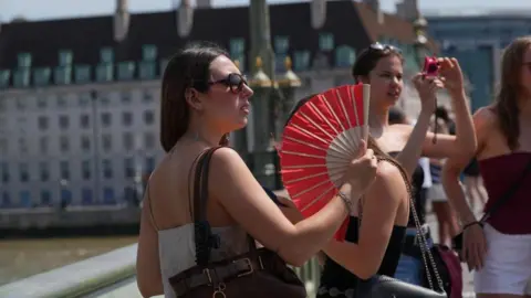 A woman with dark hair and wearing sunglasses holds a red fan to cool herself in the heat, on Westminster Bridge in central London.