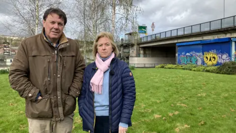 Catherine O'Sullivan and Alan O'Sullivan, Jack's parents, standing beside each other on a patch of grass under a bridge in the Hotwells area of Bristol. It is the area where the missing student was last seen.