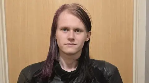 Rowan Gray Rowan Gray stands in front of a wooden door. He has black and purple long hair and he is smiling slightly. He is wearing a black biker jacket. 