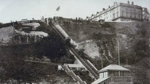 North Yorkshire Council An old black and white image of Scarborough's funicular railway. Railway tracks run vertically up a steep cliff with two buildings at the bottom and top of the track. To the left at the top of the hill, a large Victorian-era hotel can be seen.