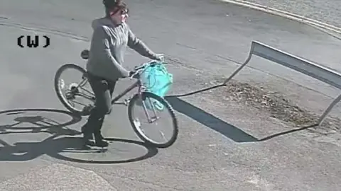 Lincolnshire Police Image of the suspect a woman with dark hair wearing a grey hooded top and dark trousers pushing a bicycle with a blue bag on the handle bars 