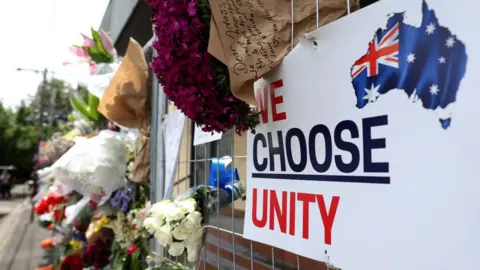 AFP Bouquets of flowers lie in a gap in a metal gate with a sign reading "we choose unity" Joined together