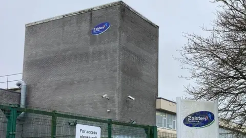 A grey building with a blue oval logo that reads 2 sisters. There is a white sign in the right foreground that also has the same logo.