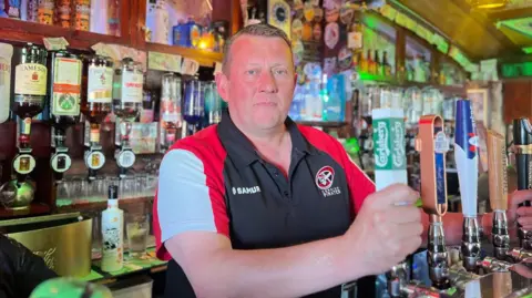 A man in a white red and black top stands behnd a bar holding a beer keg puller.