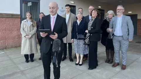 Adam Hodson lawyer for Mr Phillip's family including widow Brenda Phillips, pictured centre navy jacket, reads a statement on their behalf after the inquest. They are standing outside the court wearing formal attire.