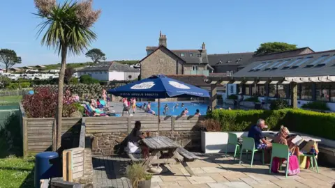 A swimming pool at a holiday park . The weather is warm and sunny and holiday makers and their children are swimming and sunbathing. Rows of caravans and motorhomes can be seen behind the pool. 