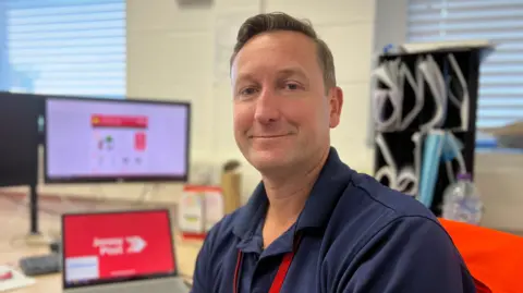 Man wearing a blue shirt with a red lanyard sat by a desk with a computer and a monitor. 