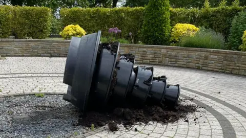 A large black plastic tiered planter has been tipped on to its side in the middle of a circular paved area of a garden. Green hedges and bushes ring the wall at the perimeter.