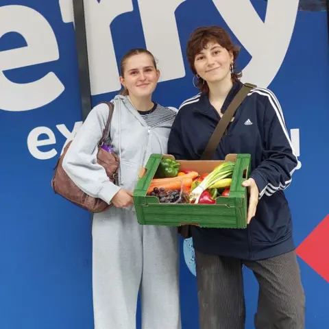 Amelie and Madeleine, two students, stand outside a Lidl store holding a box of vegetables