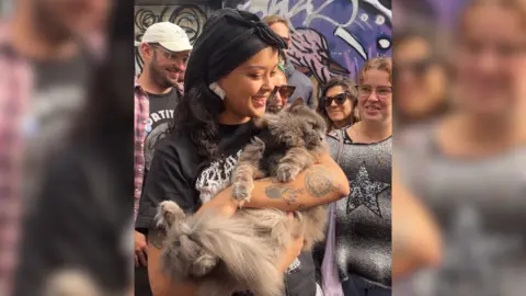Josh McLaughlin / BBC Cleo Oland cradles her cat Ashai in her arms on its back like a baby. She smiles at visitors on the street outside her home, with her cat staring at passers-by. Visitors surround Cleo and stand in front of a multi-coloured wall of graffiti art.