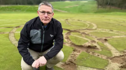 Iain Evans wearing a black jacket and tan trousers. He is crouching next to the fairway on the golf course, which has been wrecked by tyre tracks.