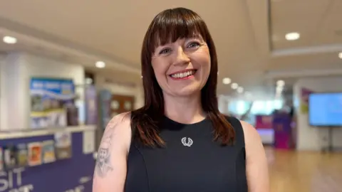 A woman smiles brightly at the camera. She has brown hair just past her shoulders with a full fringe. She wears a black dress with a white logo in the middle. Behind her is a blurred background of what appears to be a foyer.