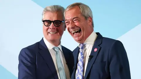 Malcolm Offord and Nigel Farage standing close together on a stage. They both have short, grey hair and are wearing blue suits and cream or white shirts. Offord has dark-rimmed glasses and is wearing a light blue tie. He is smiling while Farage, who is laughing, is wearing a paisley-patterned blue and white tie. 