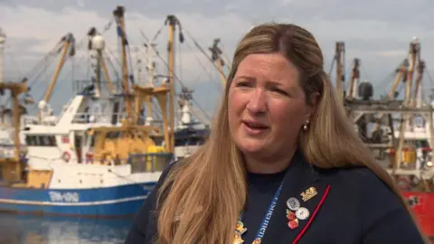 Helen Lovell-Smith is being interviewed on the quay. There are fishing boats behind her. She is wearing a collection of pin badges, two of which are Remembrance poppies.