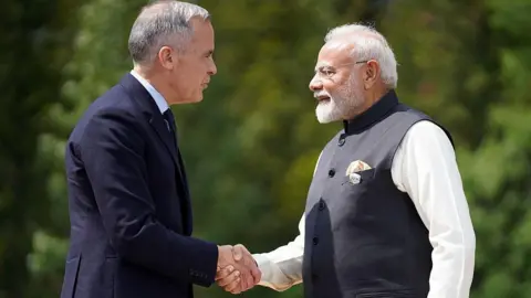 An image showing Mark Carney shaking hands with Narendra Modi, with a leafy green backdrop, in Kananaskis on the sidelines of the 2025 G7 Summit. Carney has short grey and black hair, and is wearing a black suit. Modi is wearing a grey vest with a white shirt underneath, and has short white head, a white beard and frameless eye glasses.