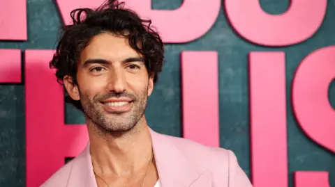 Reuters Justin Baldoni in a pink jacket, smiling and posing at the It Ends With Us premiere in 2024 in front of a dark background showing part of the film's title in giant pink letters