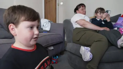 A picture of Carla, who has brown hair and is wearing a white top and green trousers, sitting in a chair with her daughter Jasmine. Her son is on the left.
