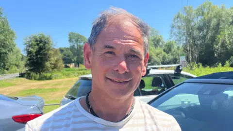 A middle-aged man faces the camera wearing a necklace and a striped T-shirt as he stands in front of cars parked on grass next to a road