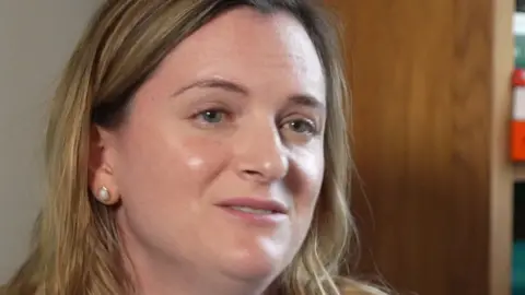 A blonde woman looks at the camera - there is a door in the background