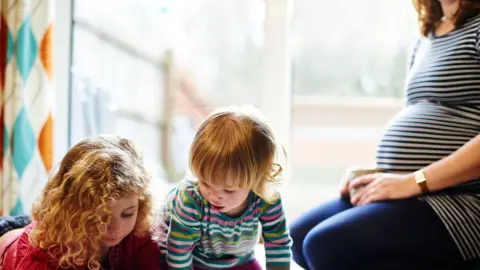 Mike Harrington/Getty Images Two young girls playing on floor of lounge while their pregnant mother watches
