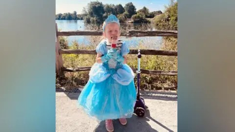 A girl in a blue dress with purple shoes holds an ice cream standing next to a scooter.