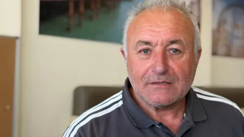 A man with short grey hair and stubble looks at the camera. He is wearing a dark grey top with white stripes on the shoulders