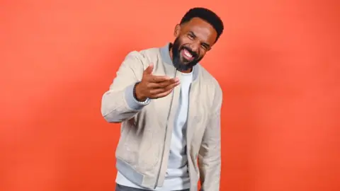 Craig David smiles and gestures to the camera - he has short black hair and a beard, and wears a cream jacket