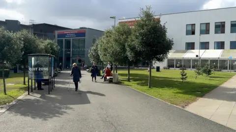 The entrance to the hospital has some trees along with path towards it with a small smoking hut on the left. A white building can be seen on the right.