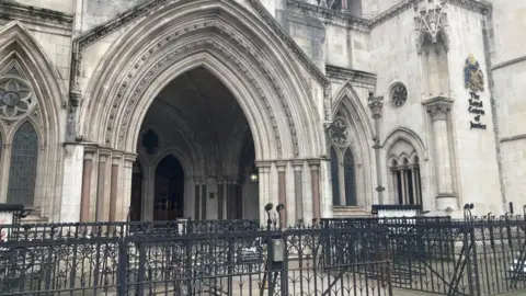 Brian Farmer/BBC A black metal fence in front of the entrance to a grey stone building. The words "The Royal Courts of Justice", in black, are mounted on a wall to the right of the entrance.