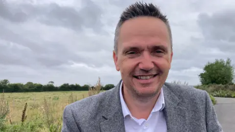 Mike Bell smiles at the camera, standing beside a small country road next to a green meadow under a grey cloudy sky. He has short greying hair and wears a grey blazer and white shirt 