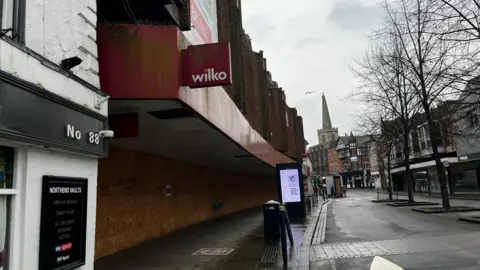 A boarded up Wilko store on a gloomy city high street, which has several empty shops. A church can be seen in the distance as a seagull flies overhead.