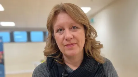 Kate Bradbrook/BBC Tracey Taylor is in the middle of the image. She has light brown shoulder length hair and is wearing a grey jumper with a black scarf. The background is blurred but two white walls, a white ceiling and a line of windows can be seen.