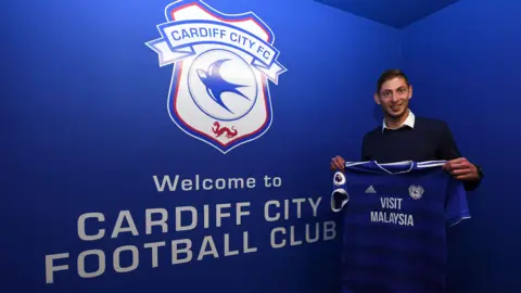 Getty Images Emiliano Sala is holding a Cardiff City jersey infront of a wall which says "Welcome to Cardiff City Football Club"