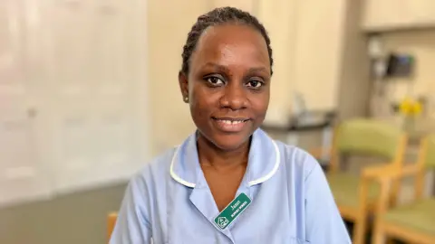 MARTIN GILES/BBC Janet, a care worker in a care home in Bedford sits on a chair and looks directly at the camera. You can see her head and shoulders. She is wearing light blue scrubs with a white trim and a green badge. She has short black hair and is smiling. You can see a few green chairs in the background. She is sitting in a training room.