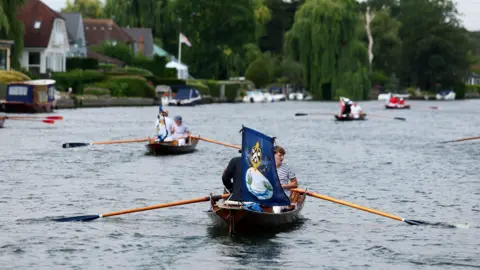 Reuters A group of traditional wooden rowing boats on a river, each crewed by individuals dressed in nautical uniform. The closest boat prominently displays a blue flag with a swan and a crown on it. The river is lined with riverside homes, boats, and greenery.