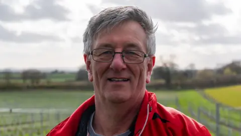Harper Adams A man with grey hair and black wire glasses is wearing a red jacket and is standing in a field that has a blurred vineyard in the background
