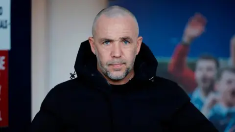 SNS Dougie Imrie - a man with a shaven head, a grey beard and moustache and a black jacket, on the touchline during a football match