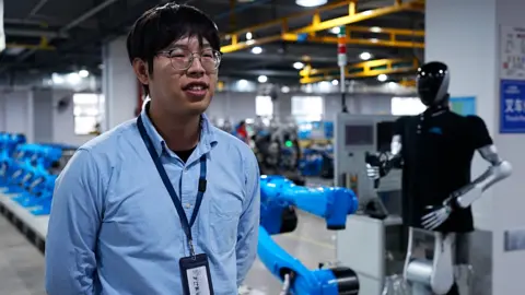 Pang Kai wearing a blue shirt with a robot standing behind him 