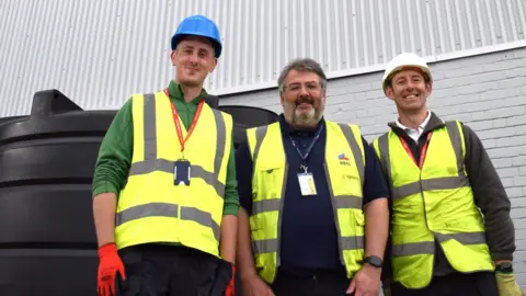 Workers at Rheinmetall BAE Systems in Telford next to the smart water butt. The three men, wearing hi-vis vests and hard hats are pictured smiling in front of a white brick building. 